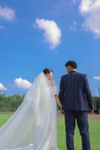 野球場で前撮り　青空の下で手を繋ぐ新郎新婦　ウェディングドレスとタキシード　