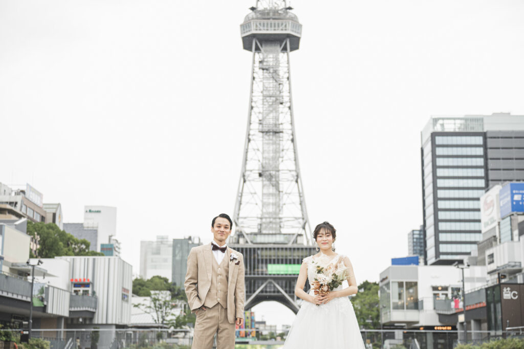 久屋大通パークで中部電力 MIRAI TOWER(旧・テレビ塔)を背景に微笑むタキシード姿の新郎とウェディングドレス姿の新婦