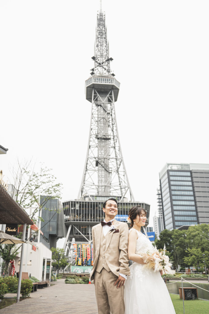 久屋大通パークで中部電力 MIRAI TOWER(旧・テレビ塔)とおしゃれなショップを背景に遠くを見つめるタキシード姿の新郎とウェディングドレス姿の新婦